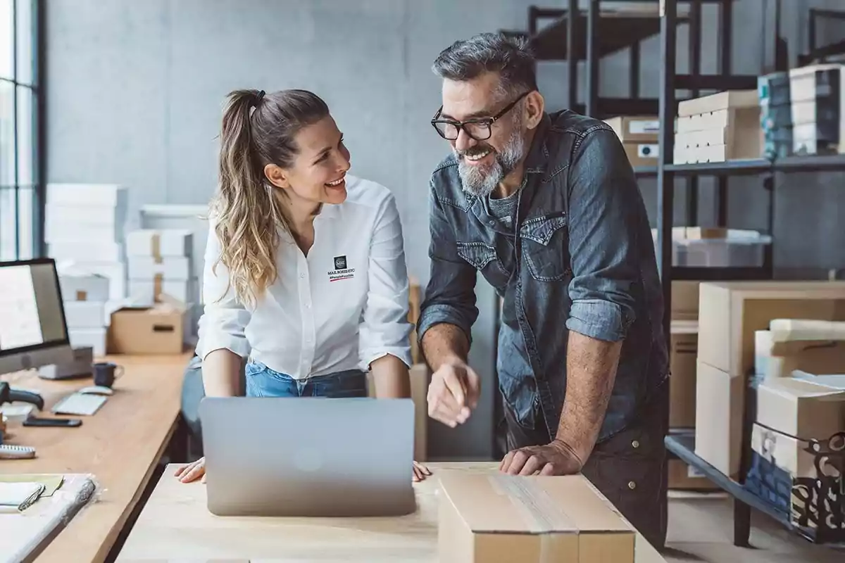 Dos personas sonríen mientras conversan en una oficina rodeada de cajas y trabajan frente a una computadora portátil