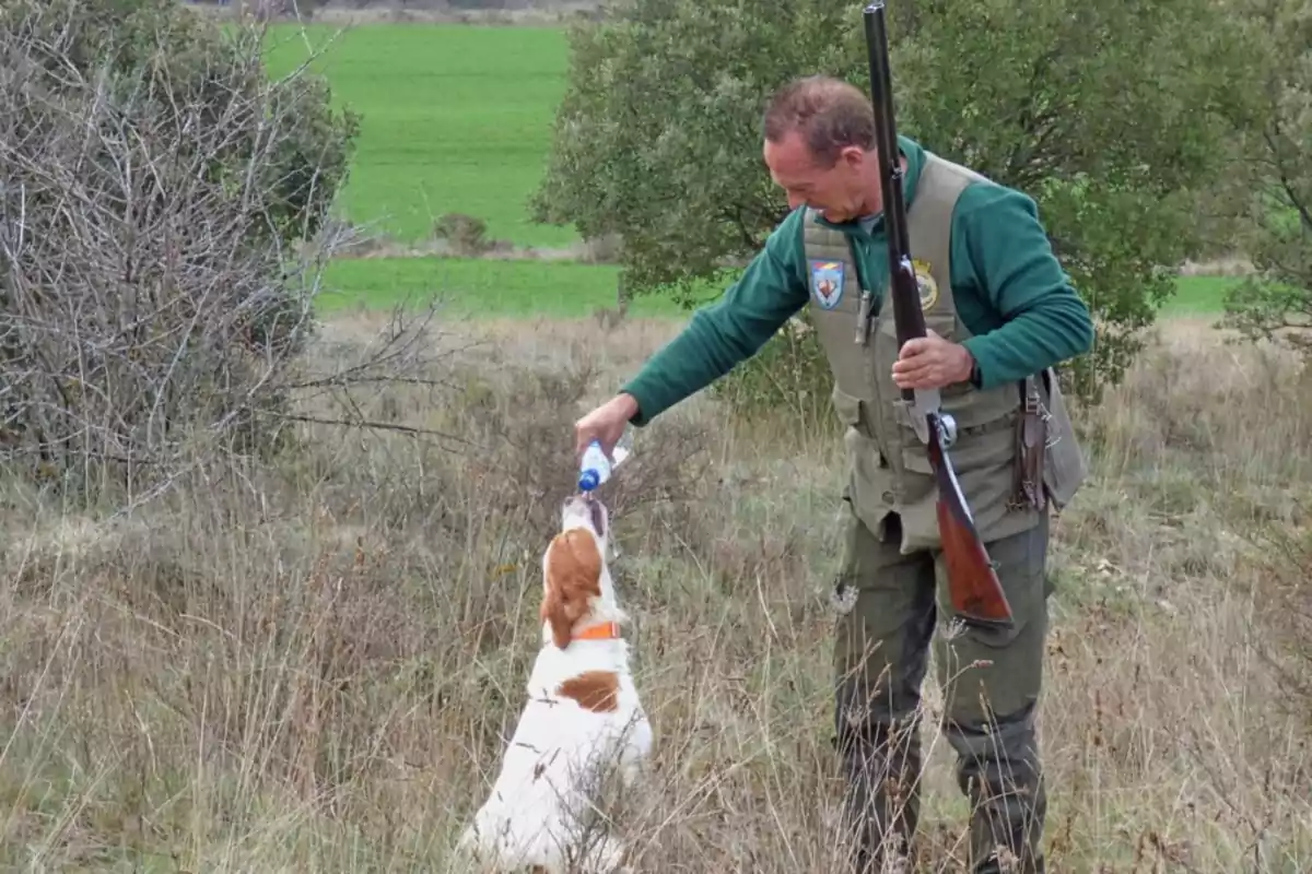 Hombre con escopeta dando agua a un perro en un campo lleno de vegetación