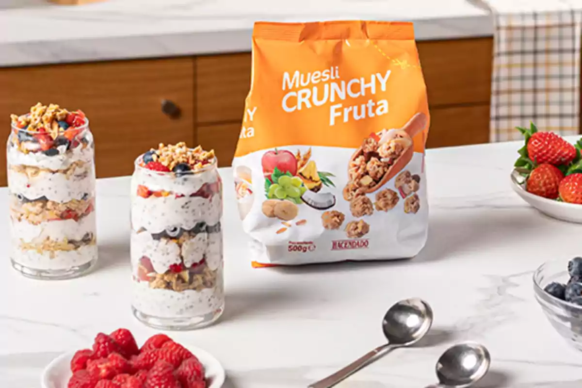 Vasos con yogur, frutas y muesli sobre una mesa junto a un paquete de muesli crunchy de fruta y platos con frutos rojos Vasos con yogur, frutas y muesli sobre una mesa junto a un paquete de muesli crunchy de fruta y platos con frutos rojos