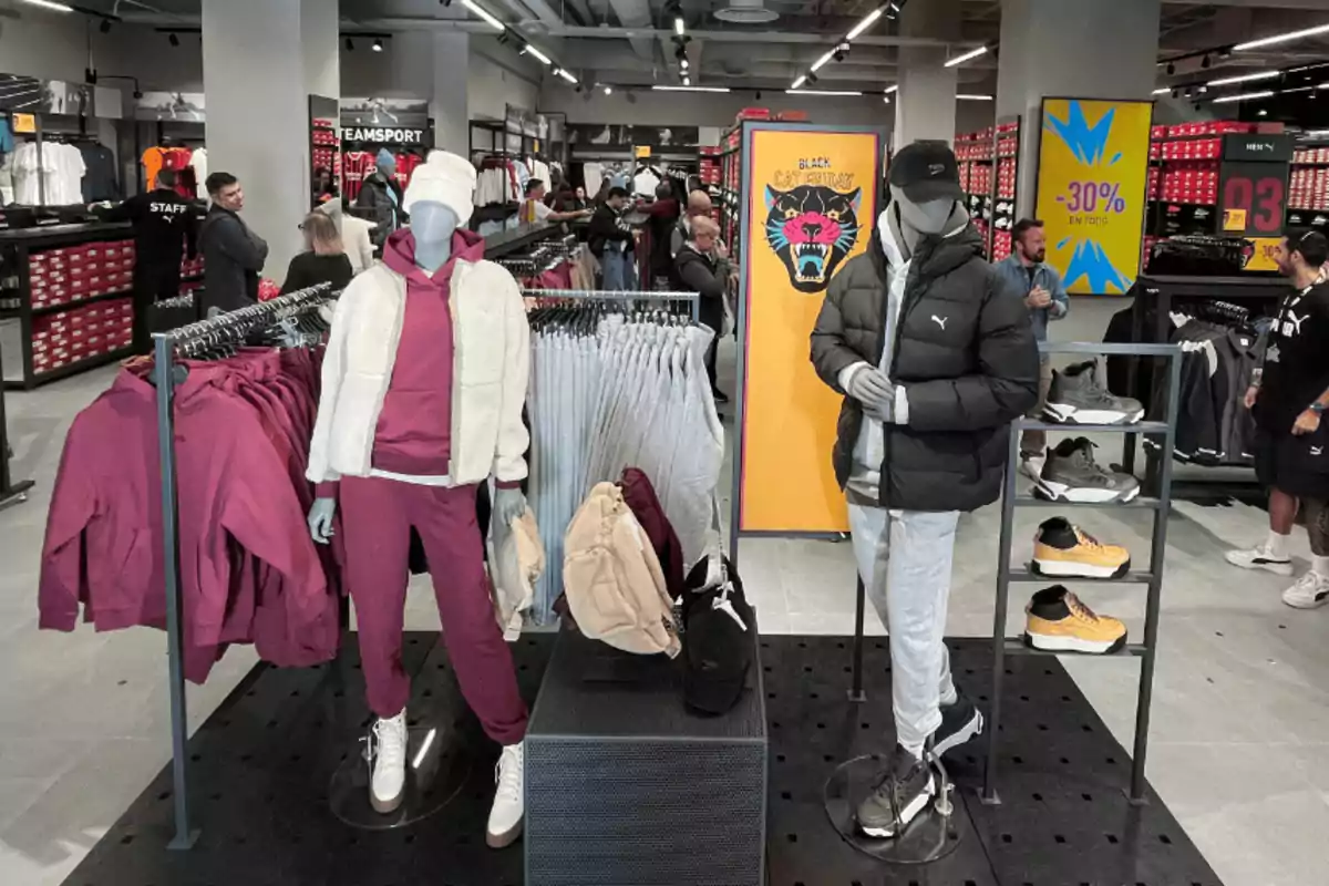 Maniquíes con ropa deportiva de invierno en una tienda de ropa con varias personas comprando y letreros de descuento en el fondo