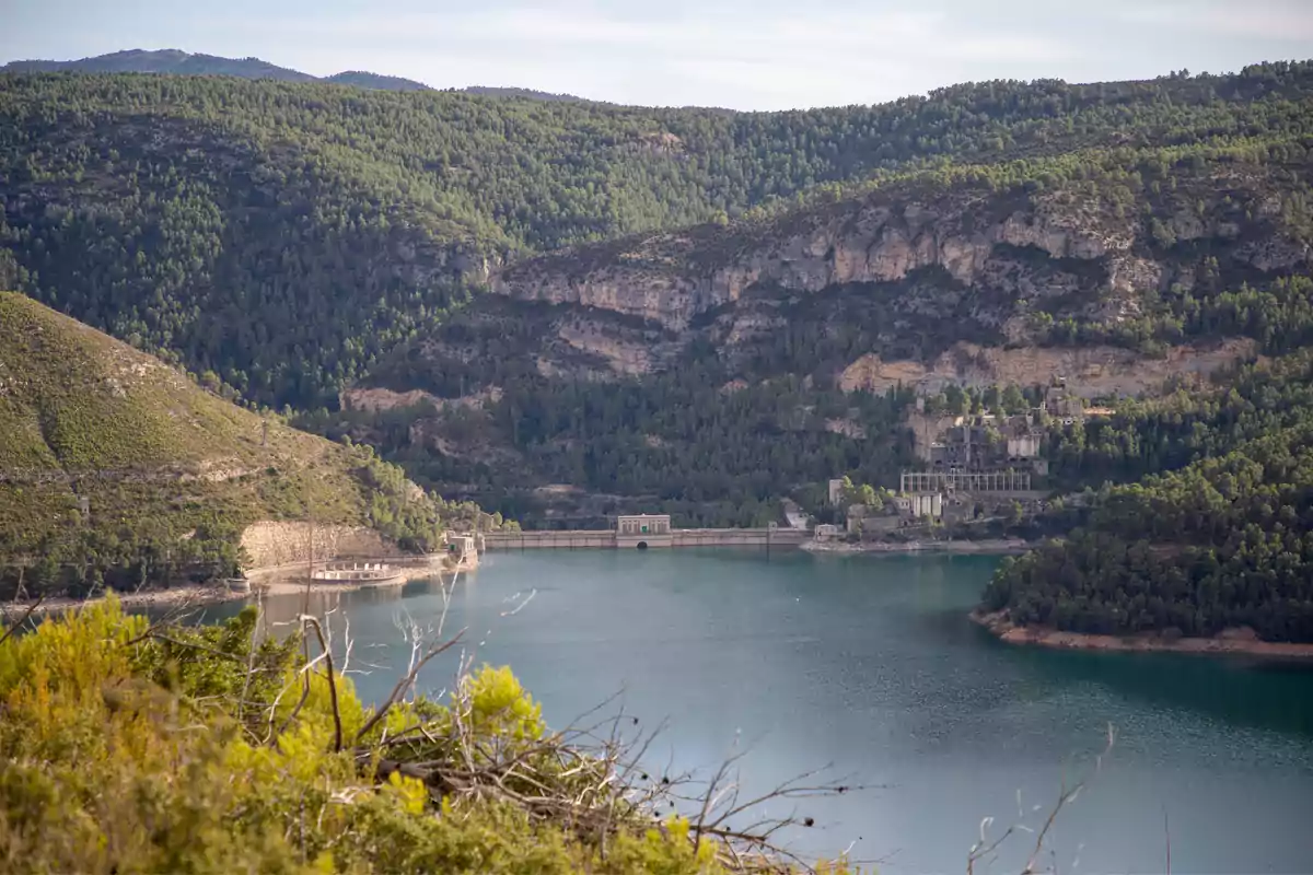Embalse rodeado de montañas cubiertas de árboles con una presa y edificaciones al fondo