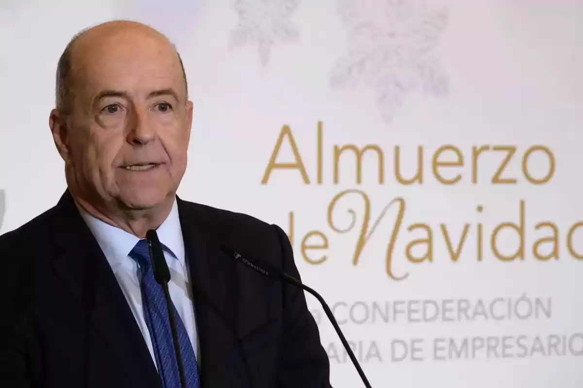 Hombre de traje y corbata azul hablando frente a un micrófono en un evento con fondo que dice Almuerzo de Navidad