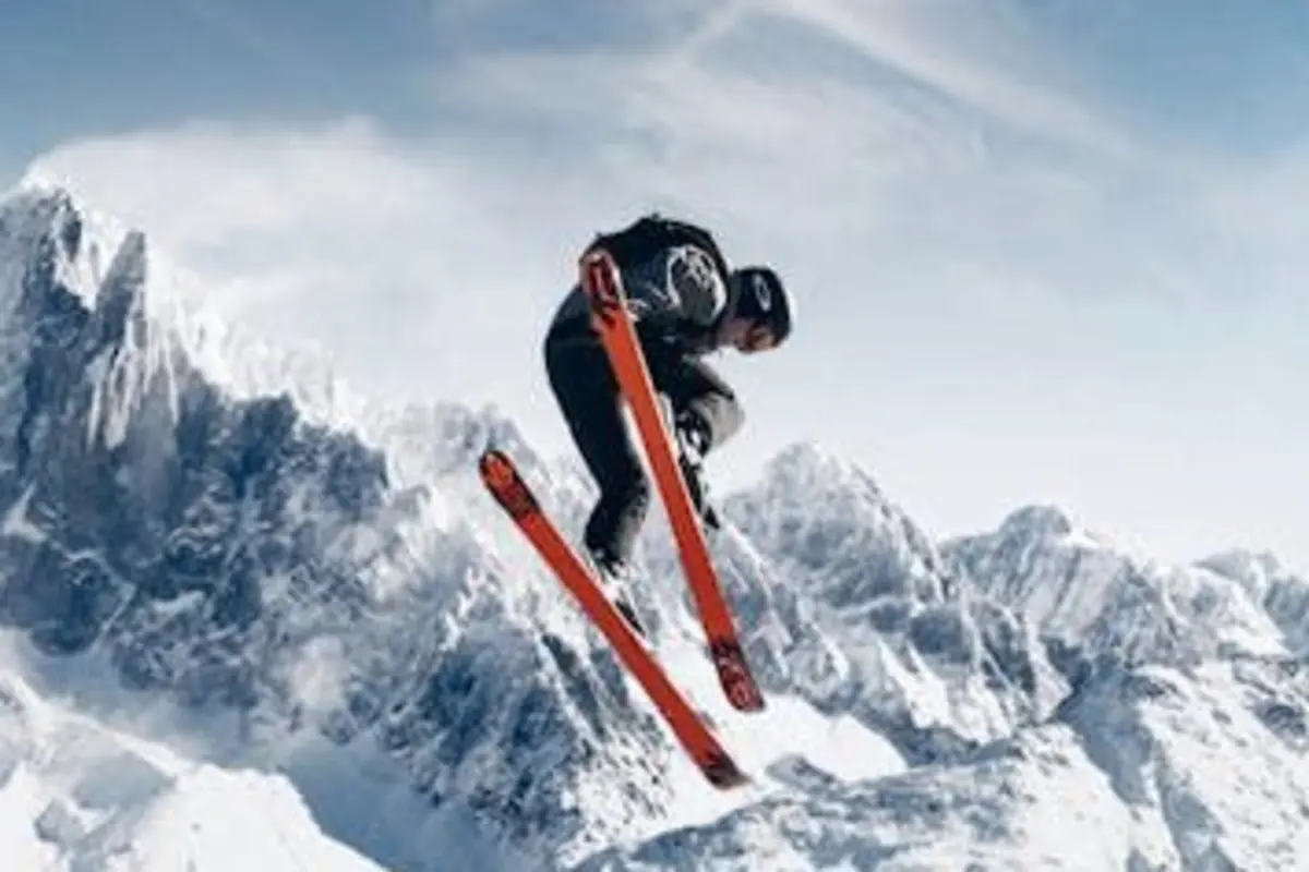 Persona esquiando en el aire sobre una montaña nevada con esquís naranjas y fondo de picos nevados