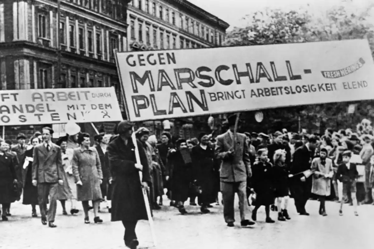 Manifestación en blanco y negro de personas marchando por la calle con pancartas en alemán en contra del Plan Marshall