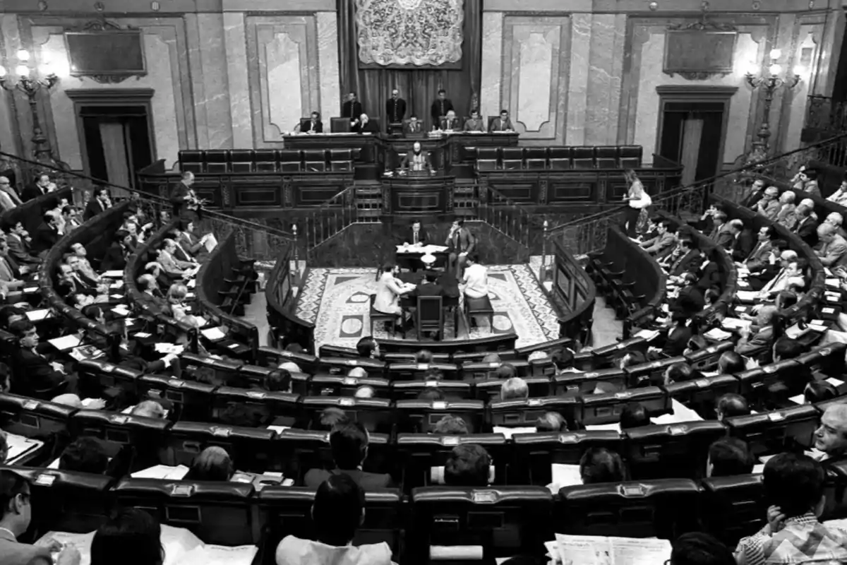 Vista en blanco y negro del interior de un parlamento con numerosos diputados sentados en sus escaños y varias personas en el centro del hemiciclo