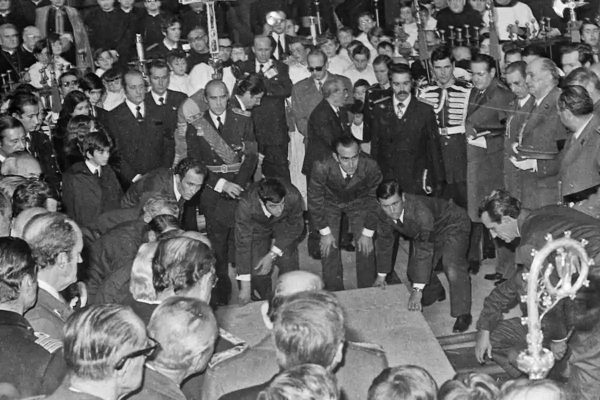 Un grupo de personas reunidas en una ceremonia solemne mientras varios hombres levantan una losa en el centro rodeados de asistentes vestidos de traje y uniforme