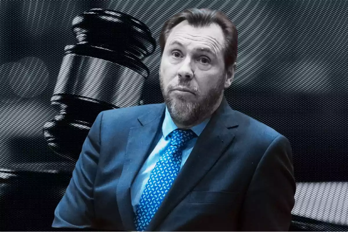 Hombre con barba y traje azul frente a un mazo de juez desenfocado en el fondo