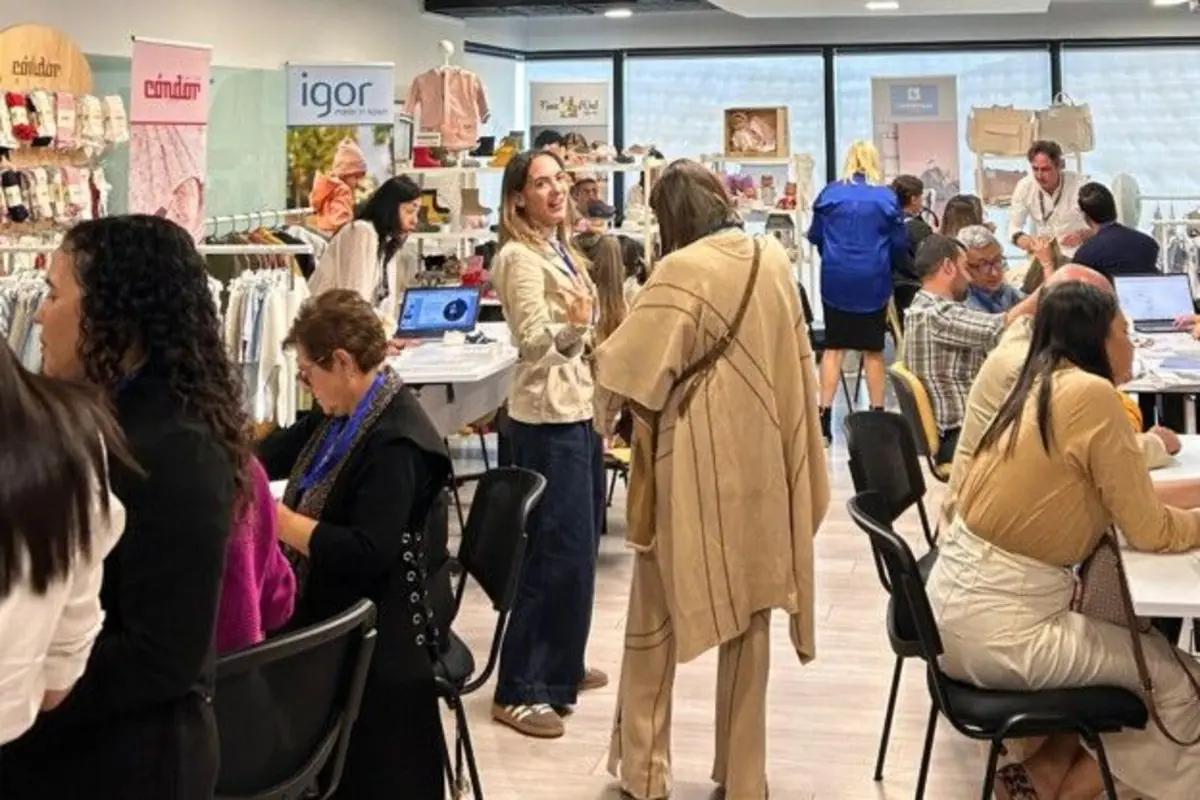 Personas conversando y trabajando en un evento de networking o feria comercial de moda infantil, con mesas, computadoras y exhibidores de ropa y accesorios en el fondo.