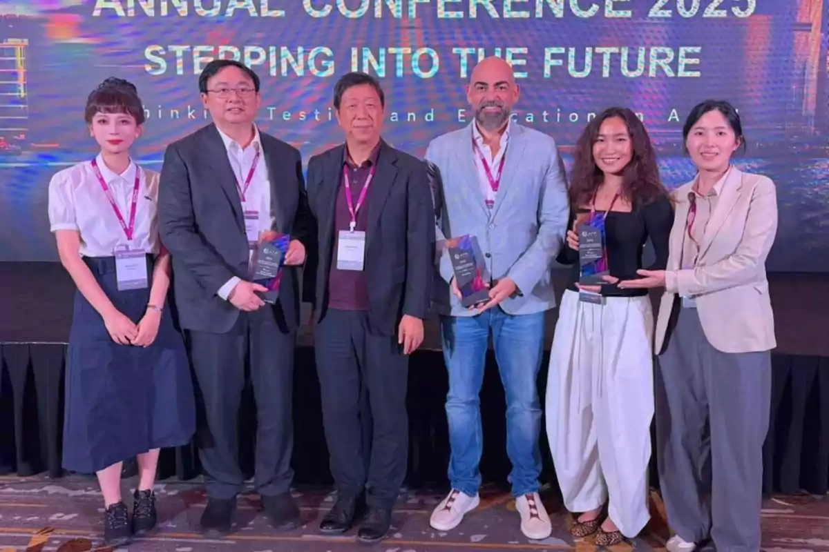 Seis personas posan sonrientes en un escenario durante una conferencia anual, tres de ellas sostienen trofeos y todos llevan gafetes de identificación.