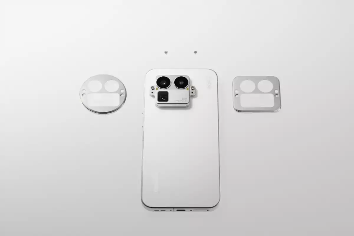 Teléfono inteligente blanco visto desde atrás con módulo de cámara grande en el centro y dos cubiertas metálicas para lentes a los lados sobre una superficie blanca