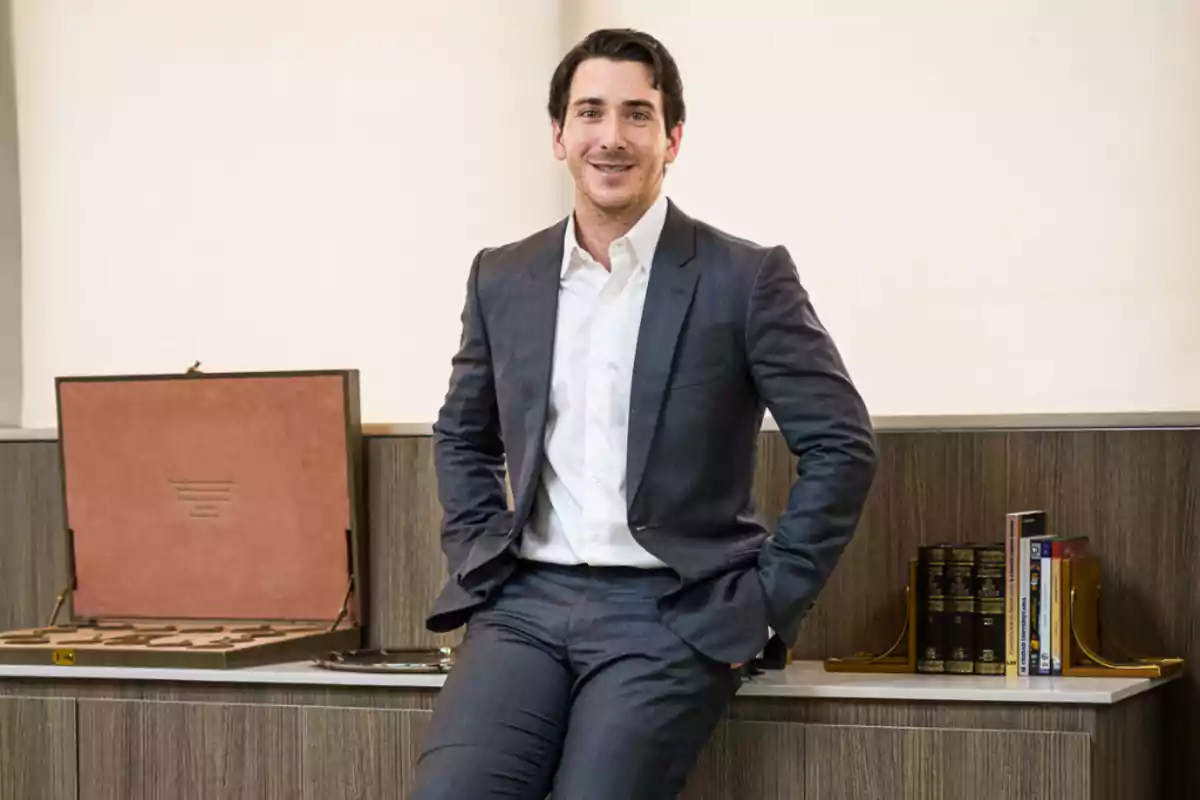 Hombre joven con traje gris y camisa blanca sentado sobre un escritorio de oficina con libros y una caja abierta a su lado