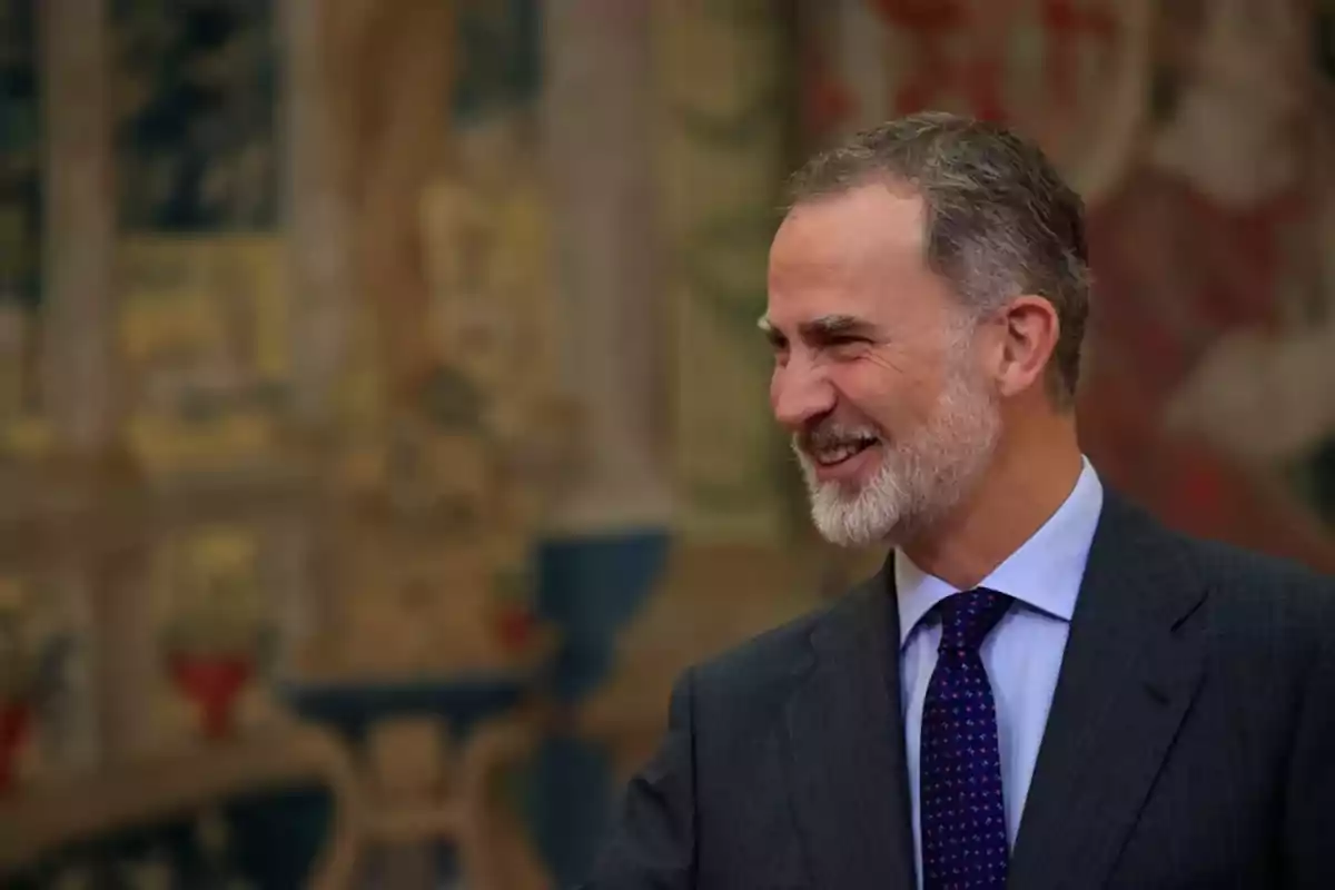 Hombre de mediana edad con barba canosa y traje oscuro sonriendo en un entorno elegante y decorado