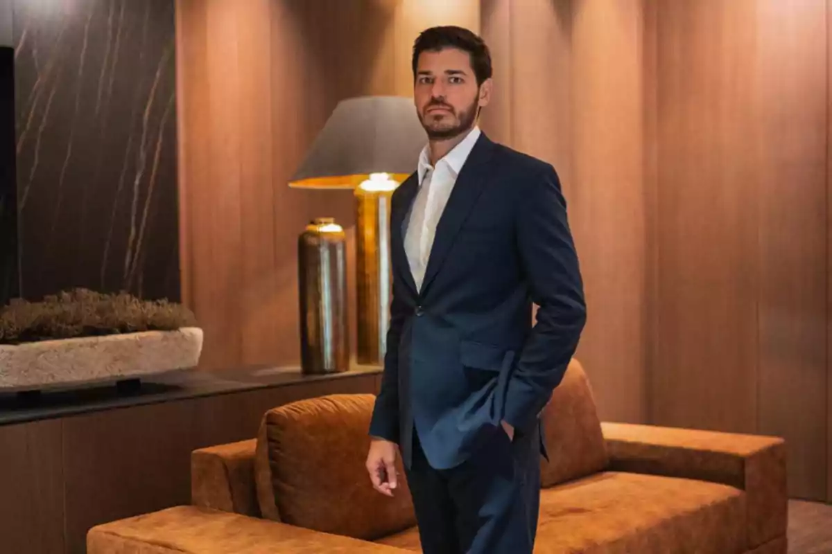 Hombre con barba y traje azul posando en una sala elegante con sofá marrón y lámpara dorada