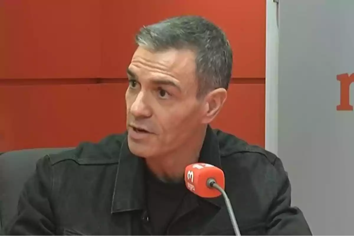 Hombre de cabello corto y canoso hablando frente a un micrófono rojo en un estudio con paredes rojas