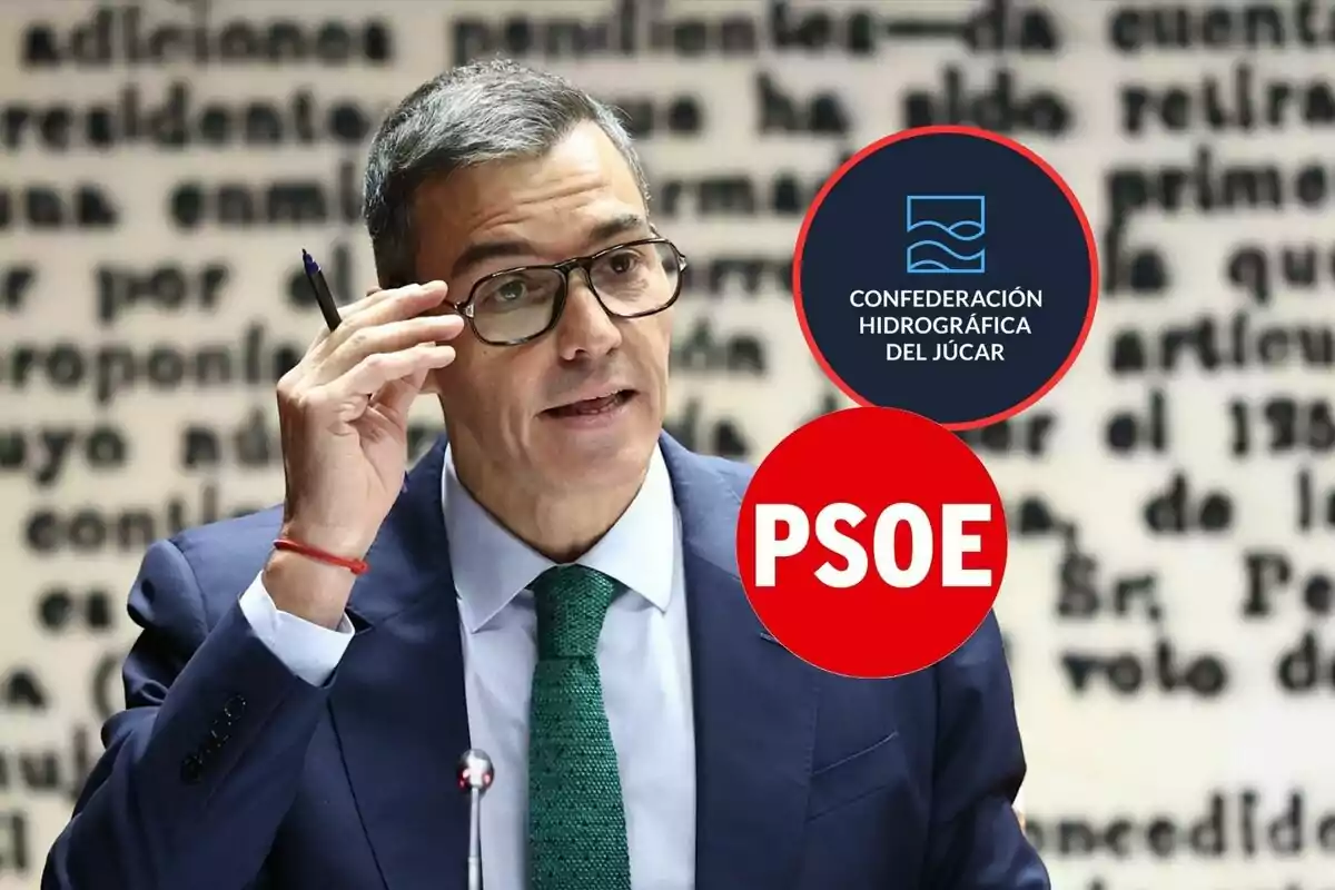 Hombre con gafas y traje azul hablando frente a un micrófono con dos logotipos superpuestos uno de la Confederación Hidrográfica del Júcar y otro del PSOE