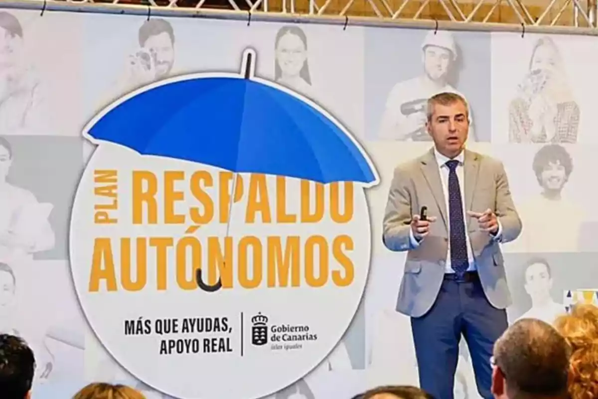 Un hombre con traje y corbata está de pie junto a un cartel grande que dice Plan Respaldo Autónomos bajo un paraguas azul, en un evento del Gobierno de Canarias