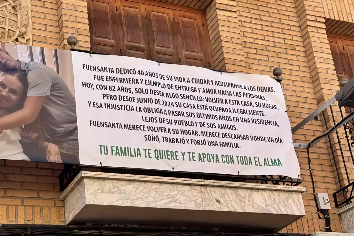 Pancarta colgada en el balcón de un edificio de ladrillo con un mensaje de apoyo a una mujer llamada Fuensanta, quien fue enfermera y ahora, a sus 82 años, no puede volver a su casa porque está ocupada ilegalmente, y su familia le expresa cariño y respaldo