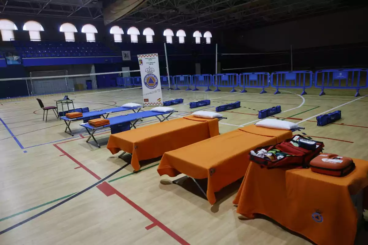 Camas plegables cubiertas con mantas naranjas y azules, mesas con equipo de primeros auxilios y un cartel de protección civil en un gimnasio adaptado como refugio temporal