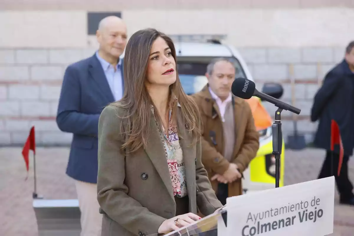 Una mujer con cabello largo y suelto habla frente a un micrófono en un evento al aire libre, con varias personas y un cartel del Ayuntamiento de Colmenar Viejo en primer plano.