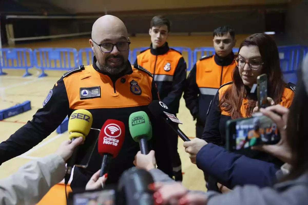 Un grupo de personas con uniformes de protección civil da declaraciones a la prensa en un recinto deportivo mientras varios micrófonos y teléfonos móviles los rodean