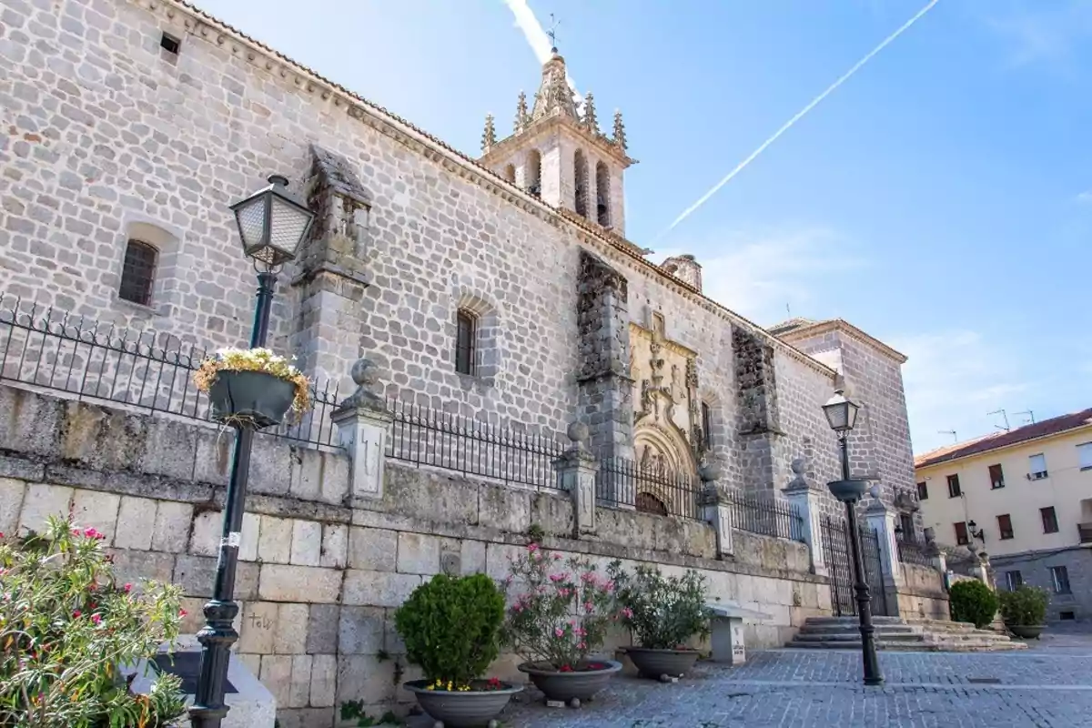 Fachada de una iglesia de piedra con una torre campanario, farolas y macetas con flores en la entrada bajo un cielo azul