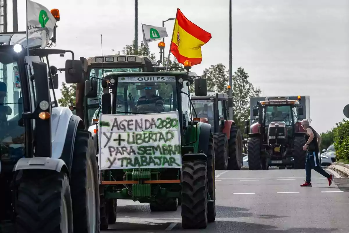 Varios tractores circulan por una carretera en una protesta agrícola, uno de ellos lleva una pancarta con mensajes reivindicativos y una bandera de España, mientras una persona cruza la calle.
