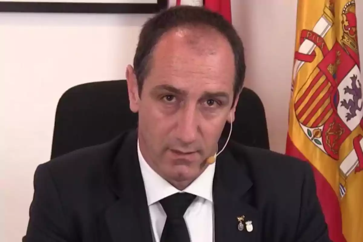 Hombre de traje oscuro con micrófono de diadema sentado frente a una bandera de España Hombre de traje oscuro con micrófono de diadema sentado frente a una bandera de España