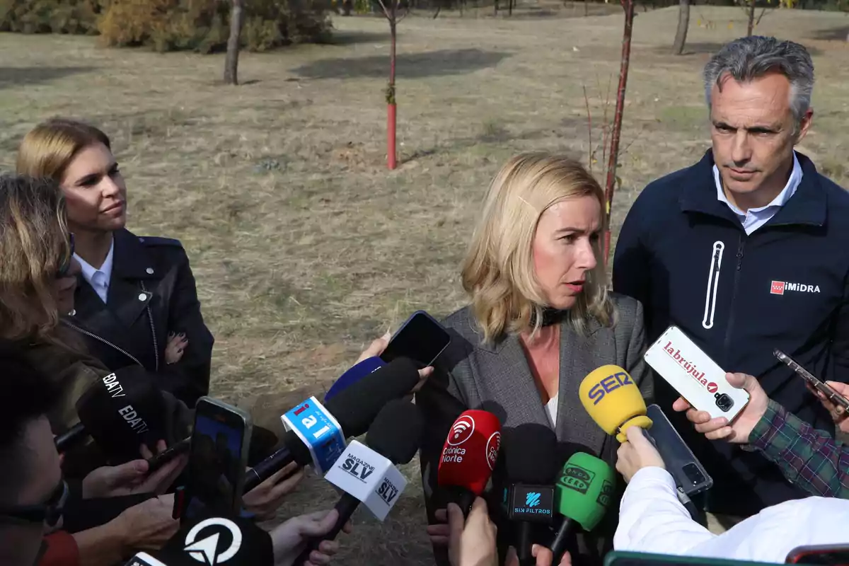 Una mujer rubia con chaqueta gris responde preguntas de varios periodistas que la rodean con micrófonos y grabadoras en un espacio al aire libre con césped y árboles al fondo
