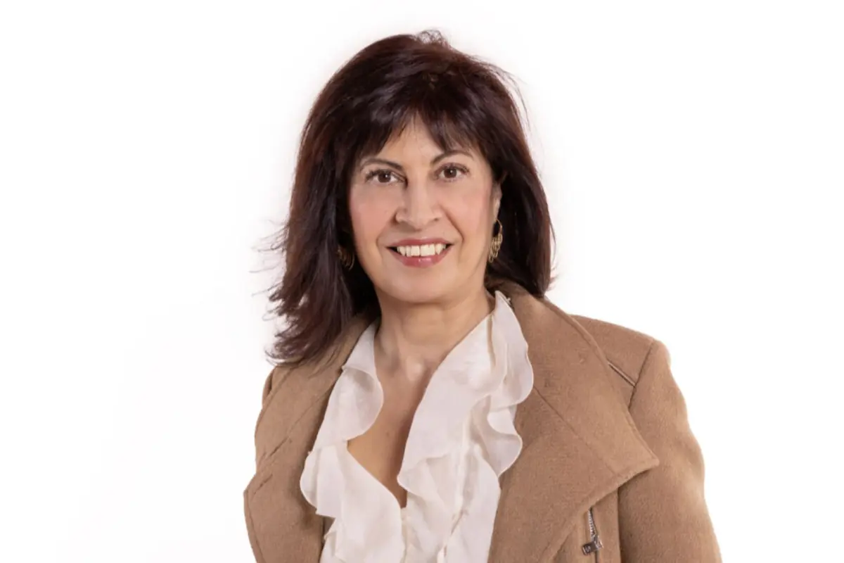 Mujer de mediana edad sonriendo con blusa blanca de volantes y abrigo beige sobre fondo blanco