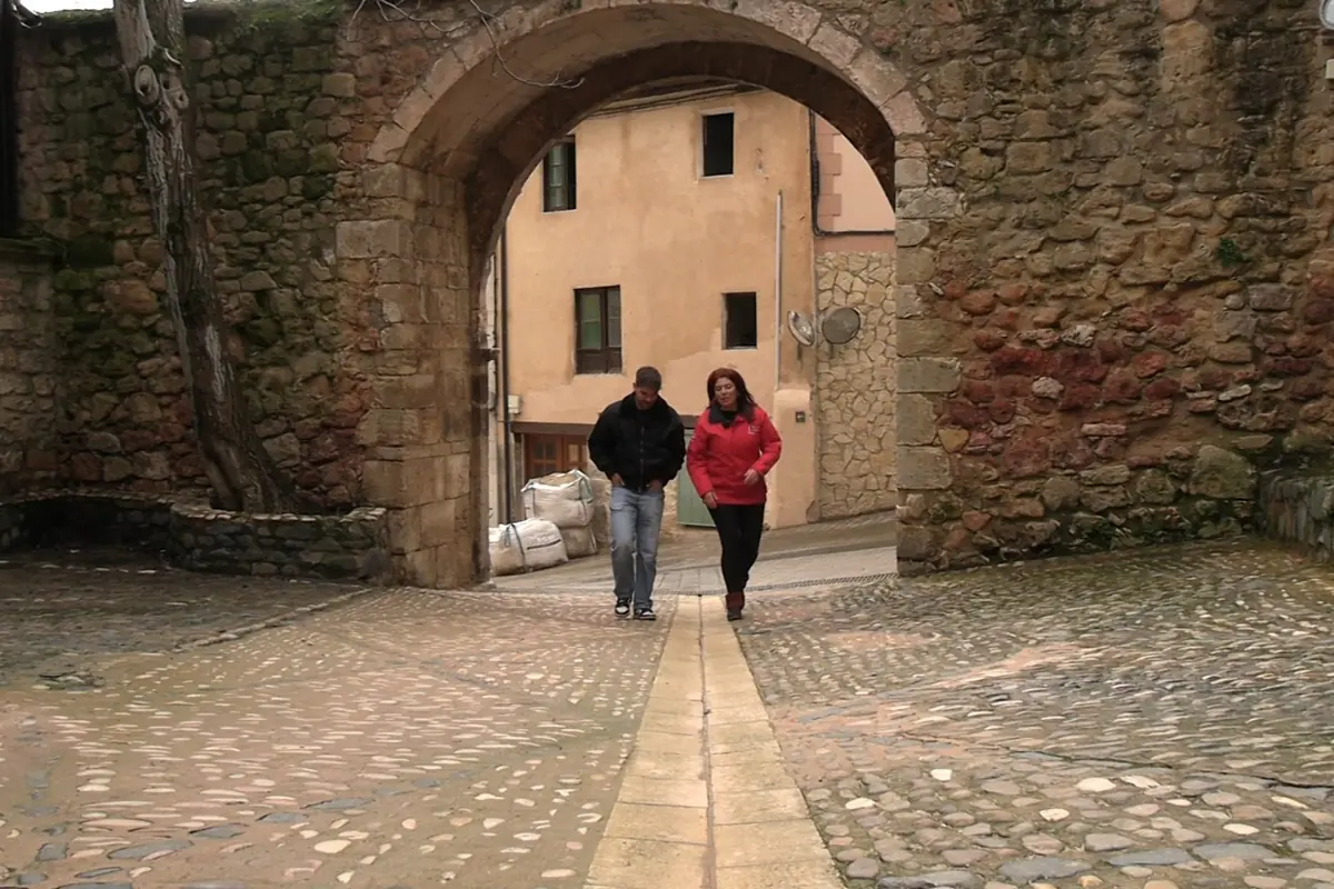 Dos personas caminando bajo un arco de piedra en una calle empedrada de un pueblo antiguo