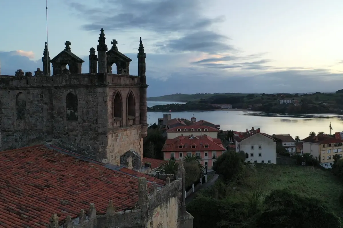 Vista aérea de una iglesia de piedra con tejado rojo junto a un pueblo costero y una ría al atardecer