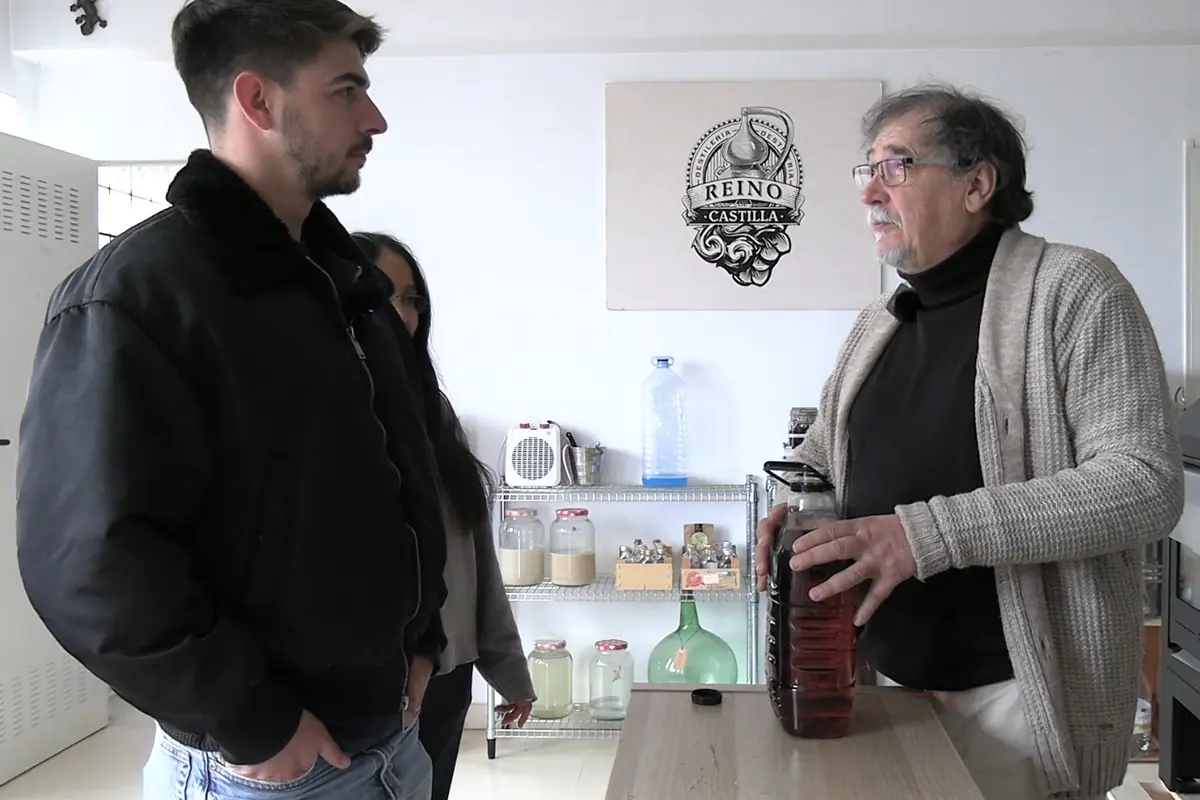 Dos hombres y una mujer conversan en una habitación iluminada, uno de ellos sostiene una botella grande con líquido oscuro sobre una mesa, al fondo hay estantes con frascos y un cartel en la pared que dice Reino Castilla