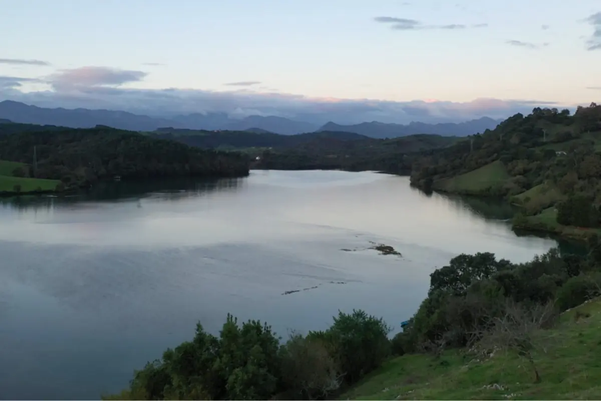Paisaje de un lago tranquilo rodeado de colinas verdes y bosques con montañas al fondo bajo un cielo despejado al atardecer