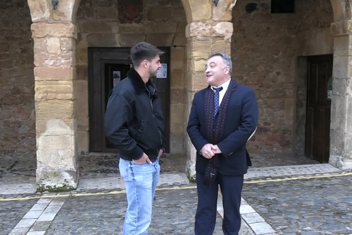Dos hombres conversan de pie en una calle empedrada frente a un edificio antiguo de piedra