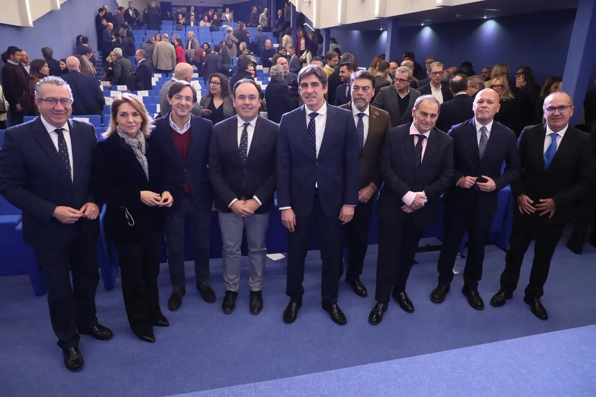 Grupo de personas vestidas de forma formal posando en la primera fila de un auditorio azul lleno de asistentes al fondo Grupo de personas vestidas de forma formal posando en la primera fila de un auditorio azul lleno de asistentes al fondo
