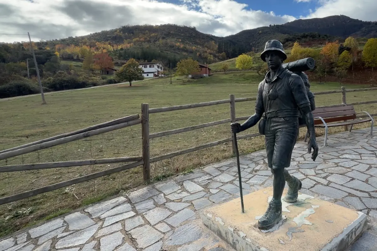 Estatua de un senderista con mochila y bastón en un mirador rural con prado casas y montañas al fondo