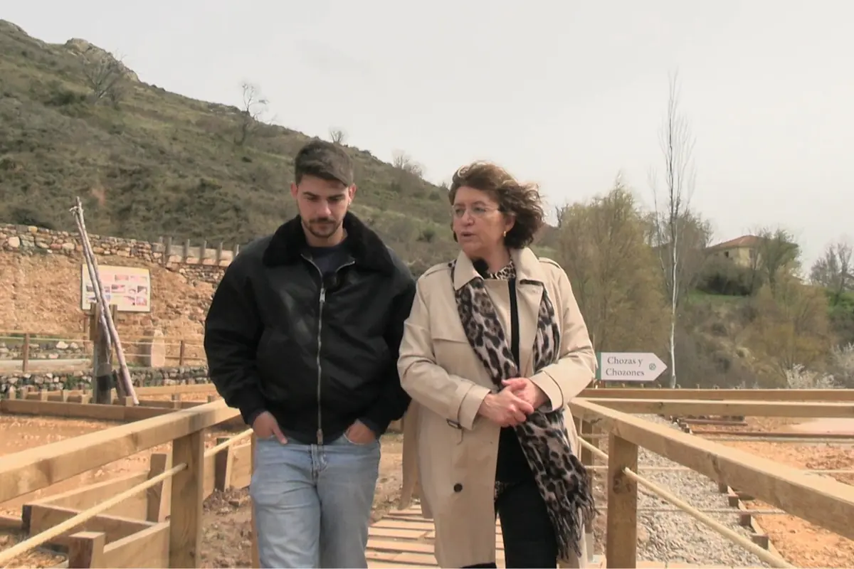 Dos personas caminando y conversando sobre una pasarela de madera en un entorno rural con colinas y vegetación al fondo