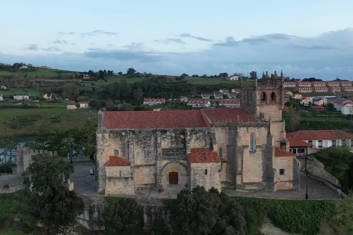 Vista aérea de una iglesia medieval de piedra con tejados rojos rodeada de vegetación y un pueblo al fondo