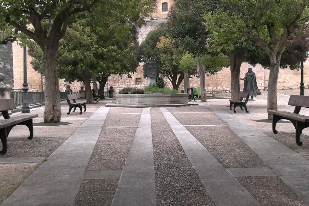 Plaza tranquila con bancos de madera, árboles y una fuente central rodeada de vegetación, al fondo una estatua y una pared de piedra