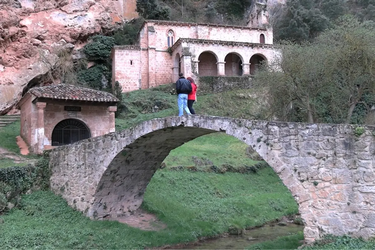Dos personas caminan sobre un puente de piedra antiguo rodeado de vegetación con un edificio histórico al fondo.