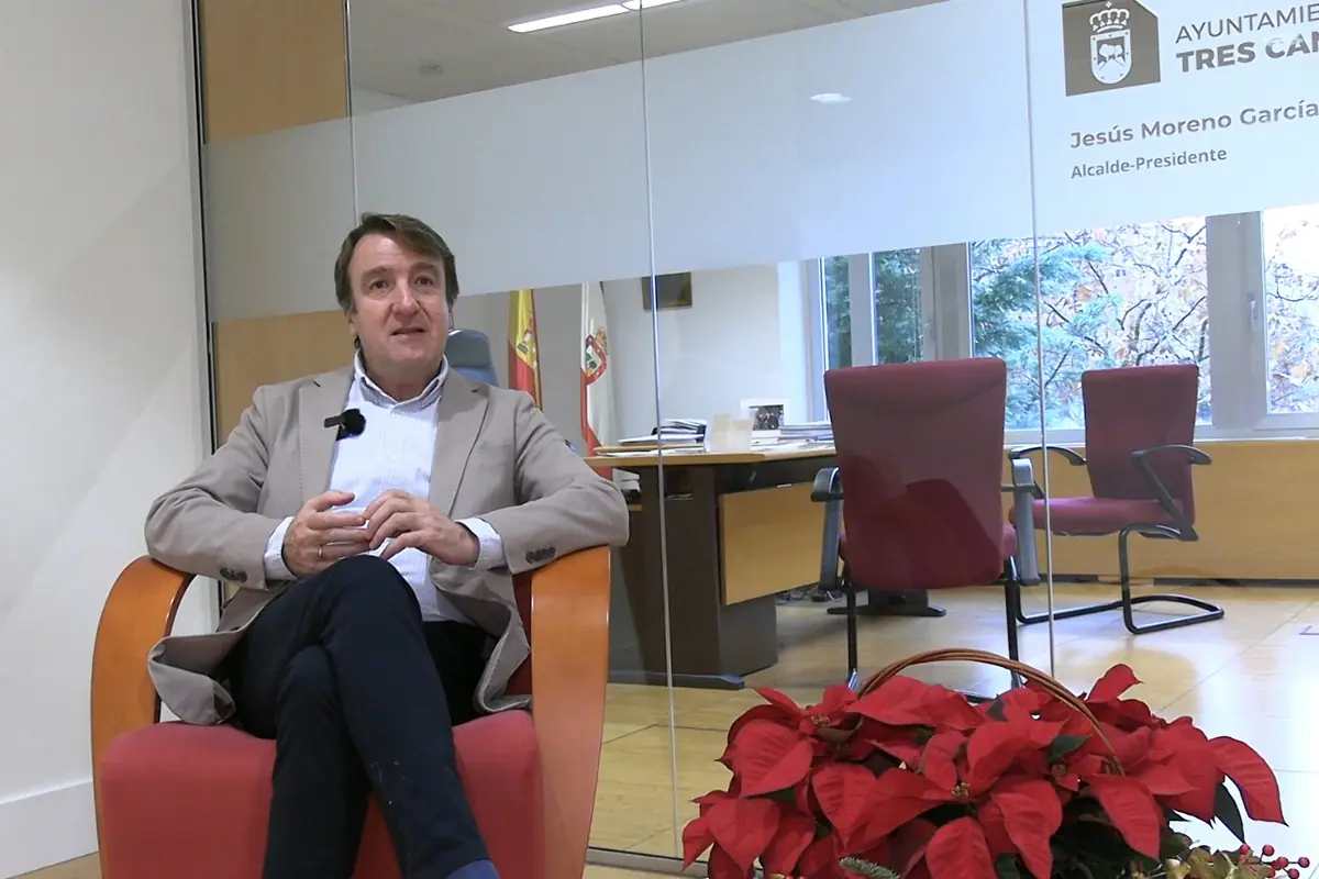 Hombre sentado en un sillón rojo dentro de una oficina moderna con cristal y rótulo institucional al fondo junto a una mesa de despacho sillas rojas y una planta de flores rojas en primer plano