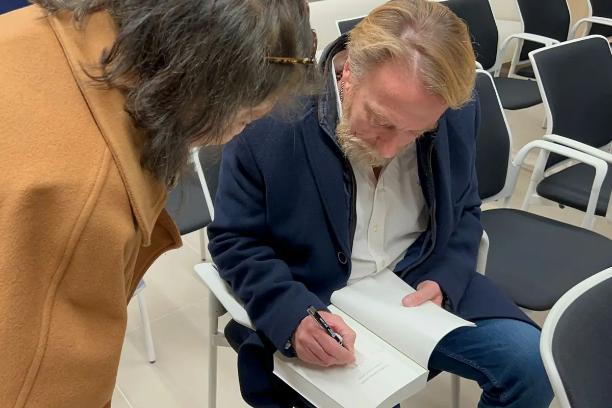 Hombre sentado firmando un libro mientras una mujer lo observa en una sala con varias sillas vacías Hombre sentado firmando un libro mientras una mujer lo observa en una sala con varias sillas vacías