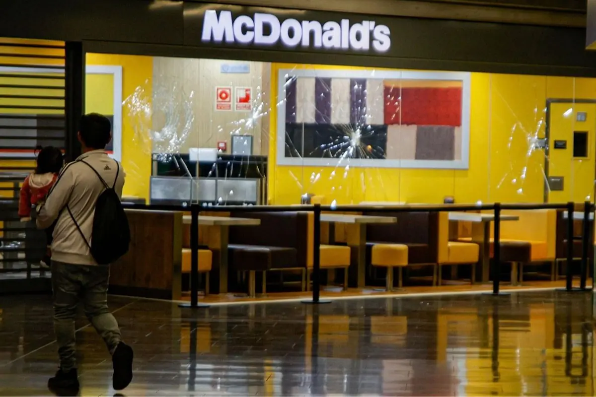 Hombre cargando a una niña camina frente a un restaurante McDonald’s vacío con vitrales rotos y mesas desocupadas Hombre cargando a una niña camina frente a un restaurante McDonald’s vacío con vitrales rotos y mesas desocupadas