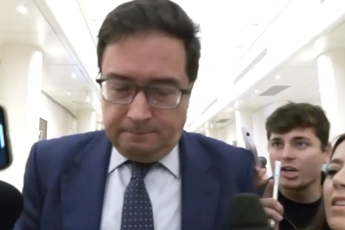 Hombre con traje azul y gafas camina cabizbajo en un pasillo mientras varios periodistas jóvenes lo rodean con micrófonos y móviles Hombre con traje azul y gafas camina cabizbajo en un pasillo mientras varios periodistas jóvenes lo rodean con micrófonos y móviles