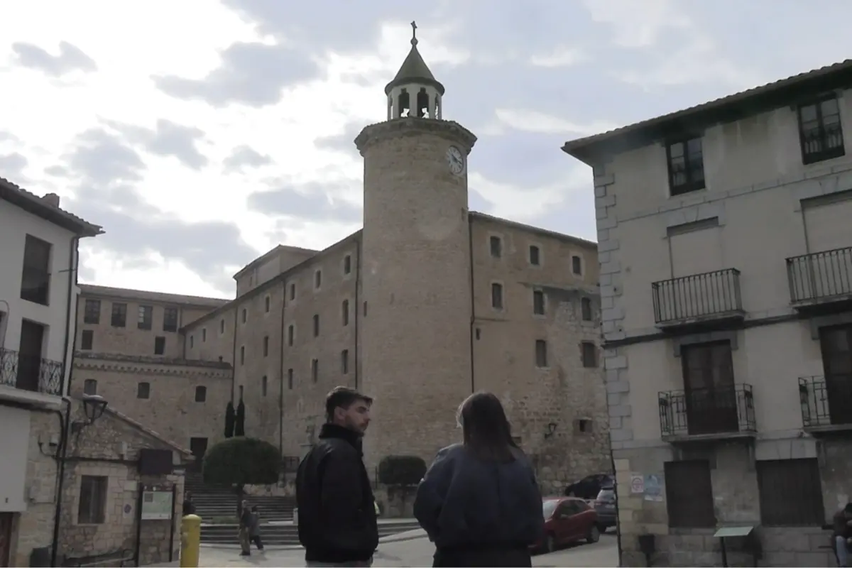 Dos personas conversan en una plaza frente a un edificio histórico de piedra con una torre y un reloj, rodeados de casas antiguas y cielo nublado.