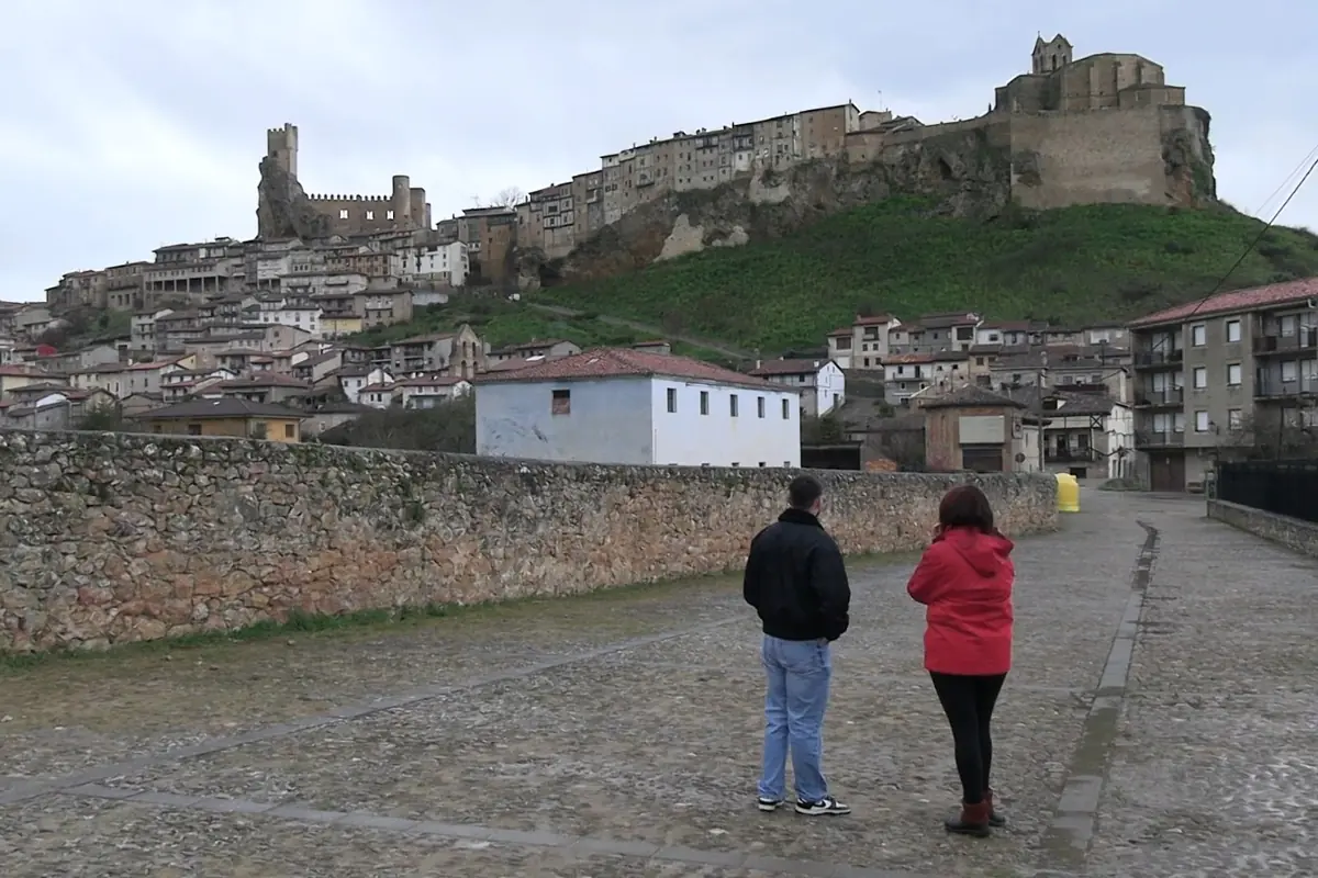 Dos personas caminan por una calle empedrada mientras observan un pueblo medieval con casas en la ladera y un castillo en lo alto de una colina