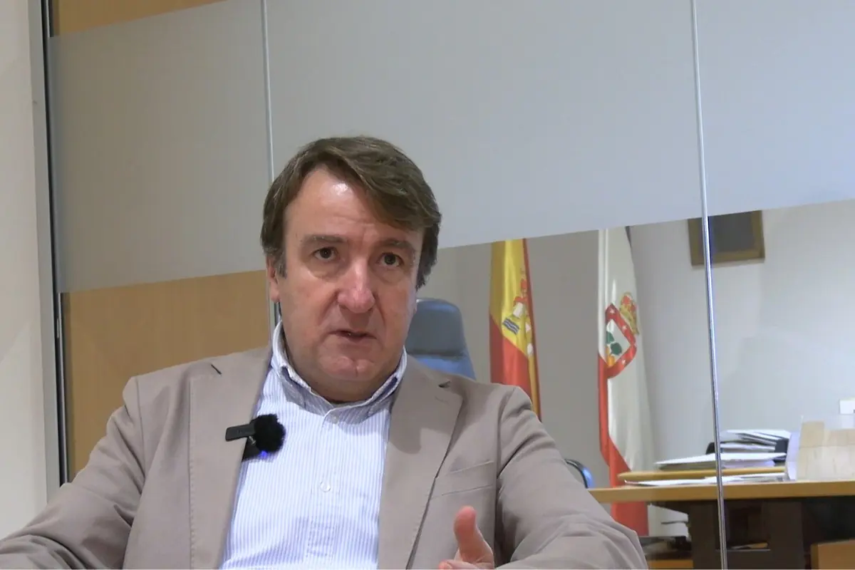 Hombre de mediana edad con chaqueta beige y micrófono de solapa hablando en una oficina con banderas españolas al fondo