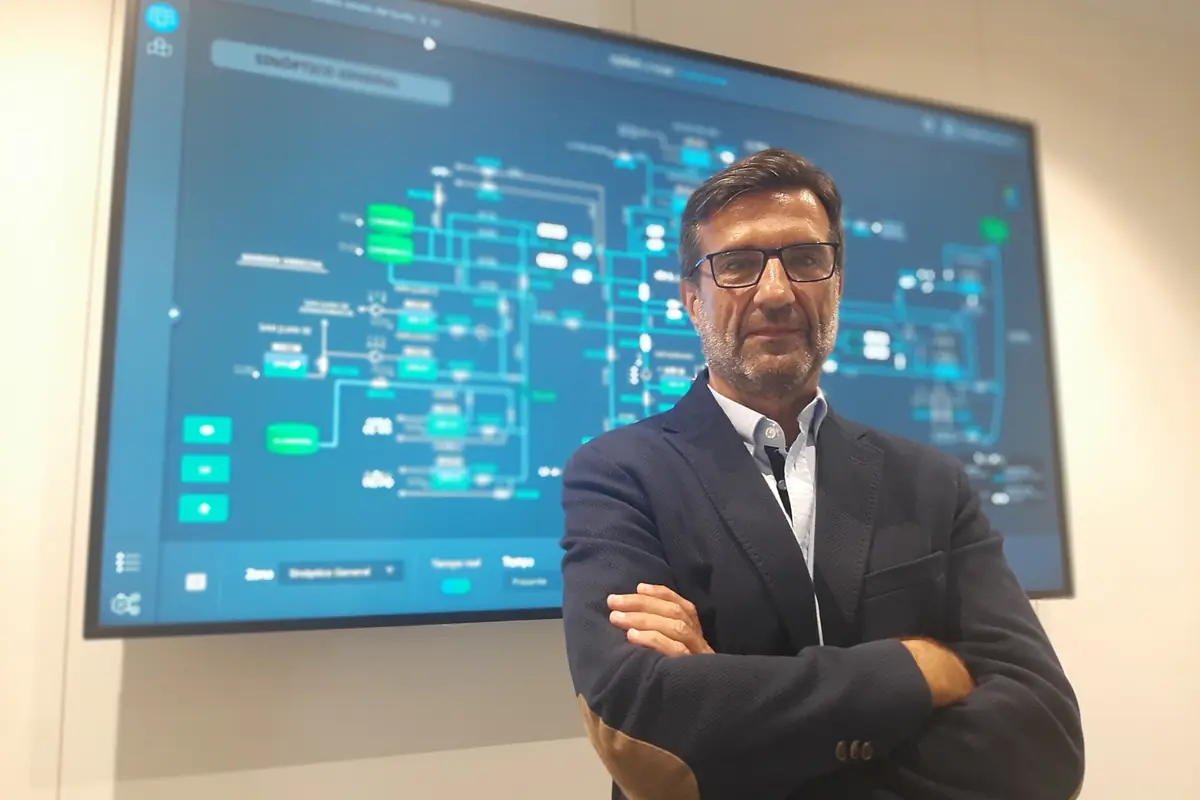 Hombre con gafas y traje cruzado de brazos frente a una gran pantalla con un diagrama tecnológico azul desenfocado