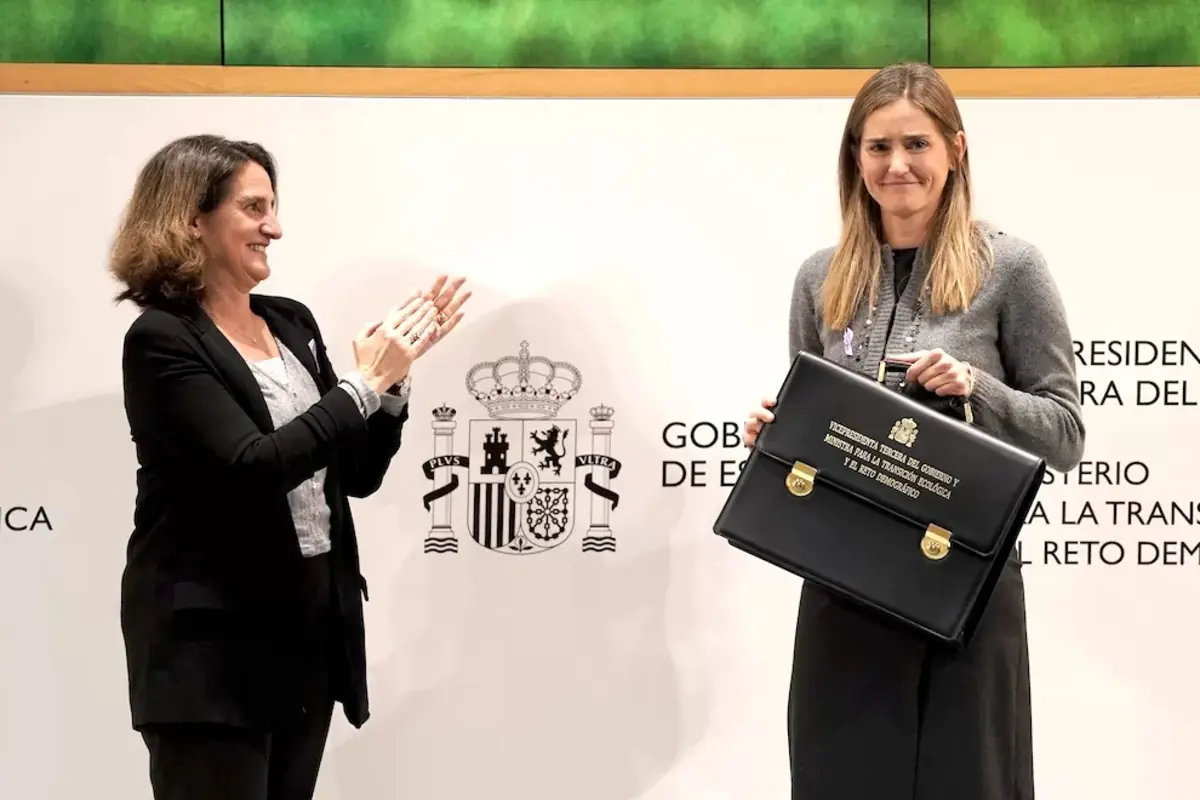 Dos mujeres en un acto oficial una de ellas sostiene una cartera ministerial negra mientras la otra aplaude frente a un fondo con el escudo de España y texto institucional