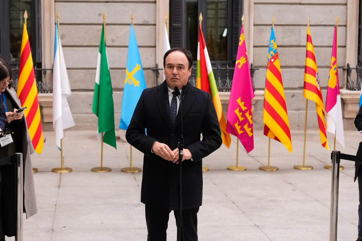 Hombre de traje oscuro hablando frente a un micrófono con varias banderas autonómicas de España al fondo en un acto oficial al aire libre